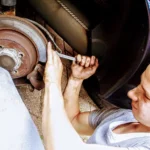 A car wheel with light gray brake dust and a mechanic pointing near the rim
