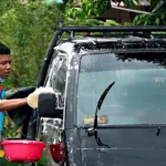 Person hand washing a silver car in a driveway with bucket and sponge in natural light