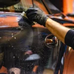 Person inspecting hard water spots on a car windshield beside a spray bottle and microfiber towel.