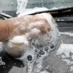 Person wiping a car windshield with a microfiber towel beside a spray bottle in natural light