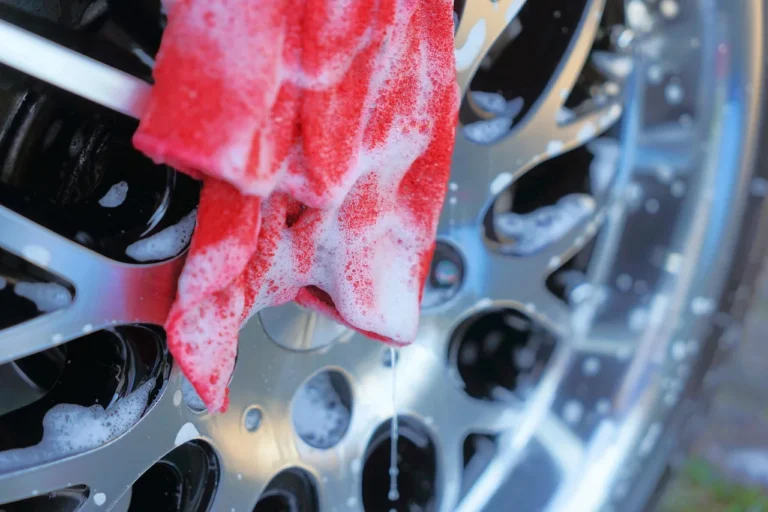 Car owner rinseless washing a dark sedan in a driveway with microfiber towels and a bucket